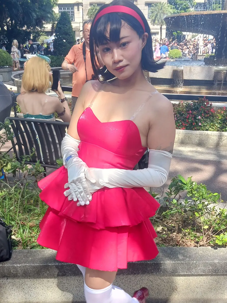 A cosplayer as Mima Kirigoe from Perfect Blue in CHAM!'s pink tiered stage dress, red headband and long white gloves, in a sunlit garden in Taipei